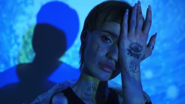 Μοντέλο με τατουάζ που καλύπτει το πρόσωπο με το χέρι σε αφηρημένο μπλε φόντο  - Πλάνα, βίντεο