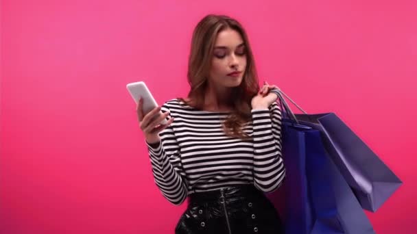 mujer reflexiva con bolsas de compras utilizando teléfono inteligente aislado en rosa - Imágenes, Vídeo