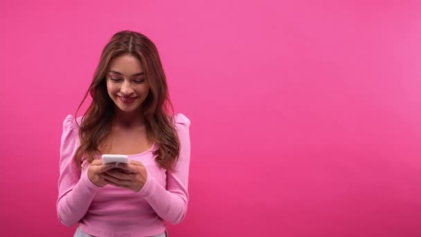 žena přemýšlí a usmívá se při chatování na smartphone izolované na růžové - Záběry, video
