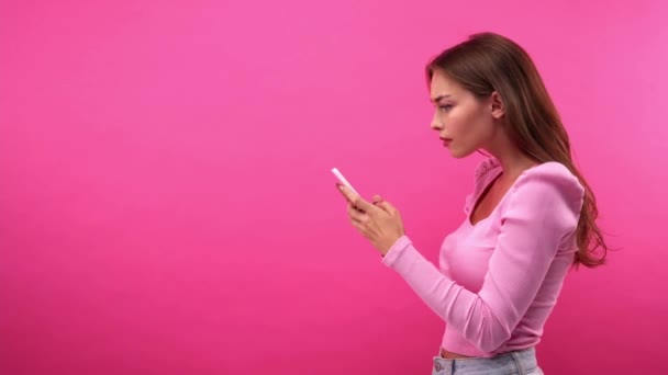 vista lateral de la mujer asombrada escribiendo en el teléfono móvil aislado en rosa - Metraje, vídeo