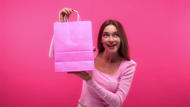 kíváncsi és boldog nő rázás bevásárló táska és gazdaság fésű elszigetelt rózsaszín - Felvétel, videó