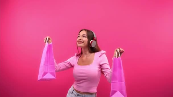 potěšená žena ve sluchátkách tanec s nákupními taškami izolované na růžové - Záběry, video