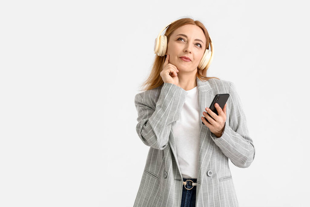 Προσεκτική ώριμη γυναίκα με κινητό τηλέφωνο ακούγοντας audiobook σε λευκό φόντο - Φωτογραφία, εικόνα