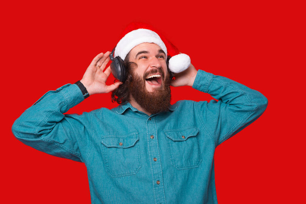 Amazed young man with beard listening music at headphones and wearing santa claus hat - Photo, image