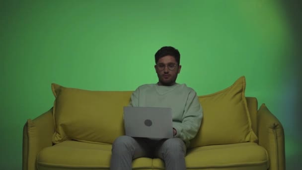 freelancer en gafas de vista sentado en el sofá y el uso de ordenador en verde - Metraje, vídeo