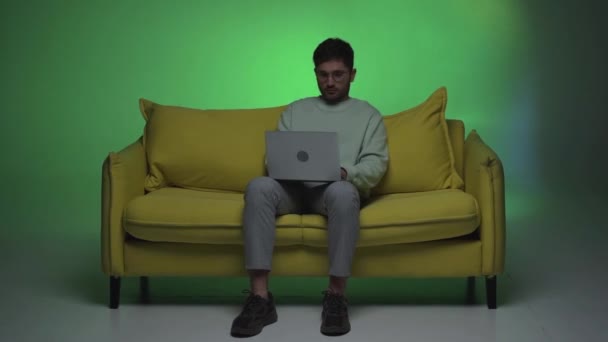 άποψη πλήρους μήκους του freelancer κάθεται στον καναπέ και πληκτρολογώντας στο laptop - Πλάνα, βίντεο