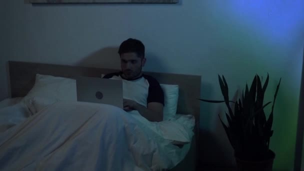 νεαρός άνδρας που εργάζεται σε φορητό υπολογιστή στο κρεβάτι το βράδυ - Πλάνα, βίντεο