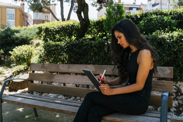 a brunette girl sitting on a bench using a tablet and talking on the phone - Foto, afbeelding