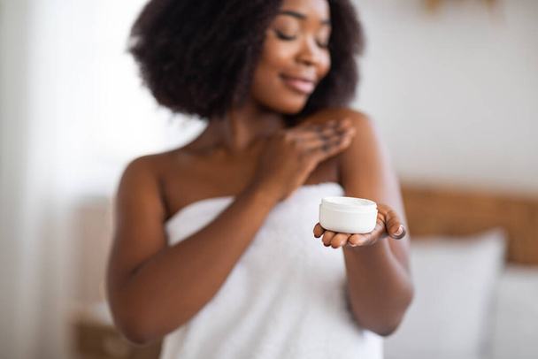 Kobiecy czarny kobieta trzyma słoik kremu do ciała lub twarzy, stosując produkt do pielęgnacji skóry po prysznicu w domu - Zdjęcie, obraz