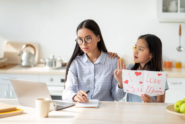 Little girl giving greeting card to her busy mother, irritated mom not paying attention, sitting in kitchen interior - Photo, Image