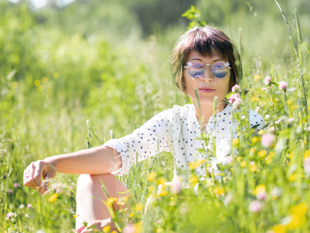 Woman in colorful sunglasses, enjoys sunlight and flower fragrance on grass field. Summer vibes. Relax outdoors. Self-soothing. - 写真・画像
