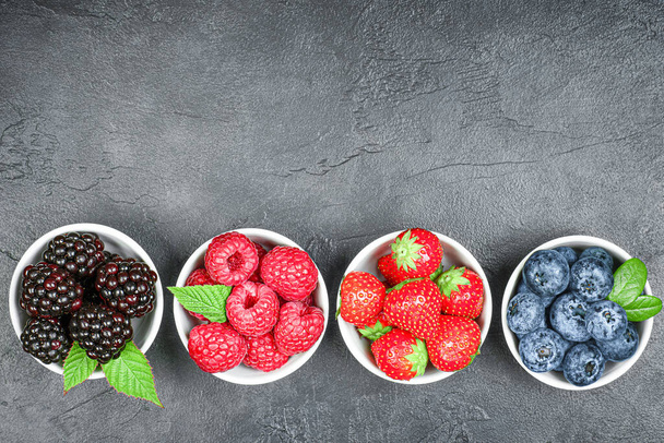 свежие ягоды малины, клубники, ежевики и черники в маленьких тарелках на бетонном темном столе - Фото, изображение