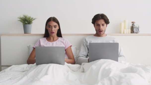 Mladí manželé pár sítí na noteboocích v posteli, dostávají e-maily a dívají se na sebe v úžasu a šoku - Záběry, video