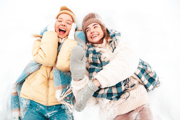Two young beautiful smiling hipster female in trendy warm clothes and scarfs.Carefree women posing in the street in park. Positive pure models lying in snow. Enjoying winter moments. Xmas concept - Fotó, kép