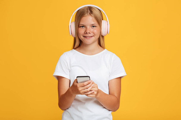 Cheerful cute teen blonde girl in headphones use phone and looks at camera - Photo, image