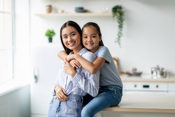 Gevoeligheid en moederschap concept. Schattig aziatisch meisje knuffelen moeder van de achterkant, zitten op diner tafel in keuken - Foto, afbeelding