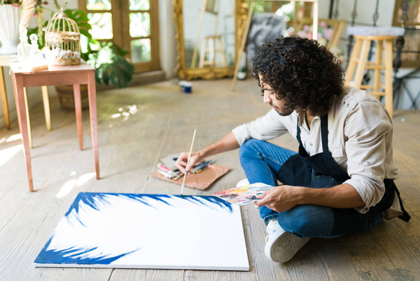 Ισπανόφωνος νεαρός κάθεται στο πάτωμα του στούντιο και αρχίζει να ζωγραφίζει ένα έργο τέχνης σε ένα λευκό καμβά - Φωτογραφία, εικόνα