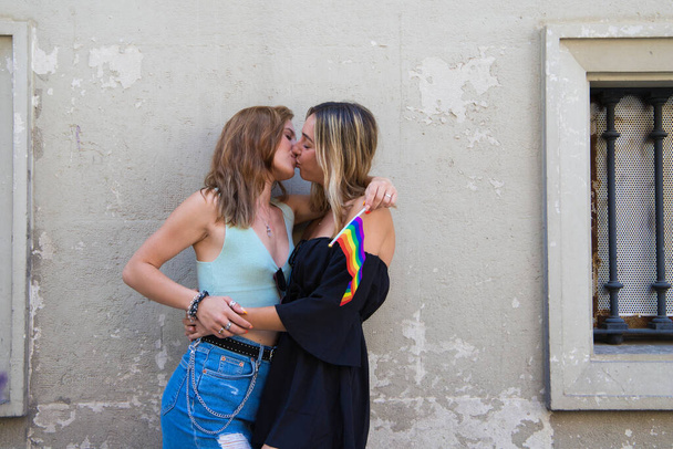 Красивые молодые женщины обнимают друг друга с флагом гей-гордости в руках. Девочки лесбиянки и страстно целуются. Концепция равенства и разнообразия. - Фото, изображение