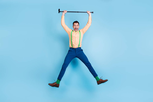Foto von energischen Kerl springen halten Gehstock heben Arme tragen Hosenträger Hemd Fliege isoliert blaue Farbe Hintergrund - Foto, Bild