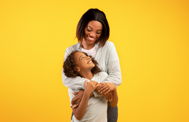 Mothers love and care. Happy young african american mom embracing her cute little daughter, yellow background - Photo, image