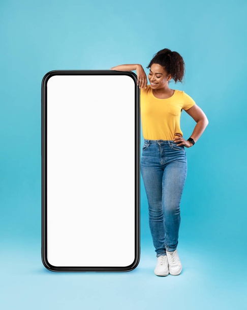 Joyful Afro woman in casual wear leaning on huge cellphone with empty white screen on blue background, mockup - Photo, image