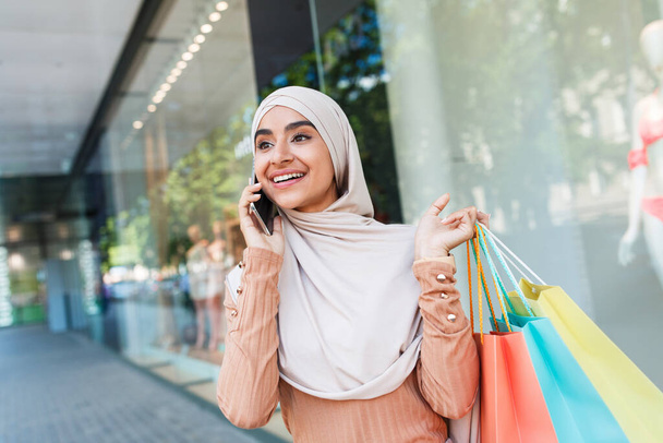 Ευτυχισμένος χαριτωμένο νεαρή γυναίκα Άραβας σε hijab ματιά αγγελία για την τεράστια πώληση, απολαμβάνει μοντέρνα ψώνια με πολλά πακέτα - Φωτογραφία, εικόνα