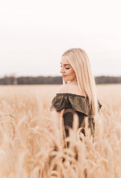 Μια νεαρή όμορφη ξανθιά γυναίκα με μακριά μαλλιά περπατά μέσα από ένα χωράφι με σιτάρι το καλοκαίρι - Φωτογραφία, εικόνα
