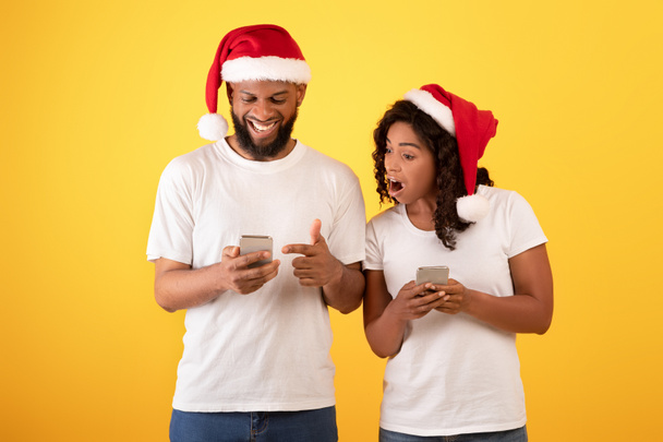 Wow Christmas mobile app. Surprised black spouses looking at smartphones, posing and wearing Santa hats - Foto, afbeelding