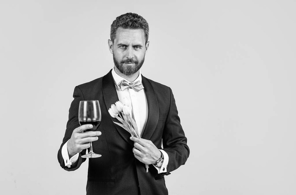 handsome tuxedo man in bow tie with flower bouquet greeting with glass of red wine, happy valentines day, copy space - 写真・画像