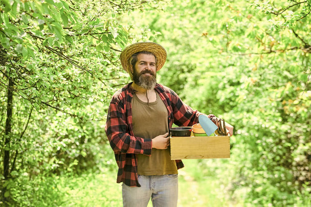 eco farm. mature man gardener working in greenhouse. brutal male carry garden tools. human and nature. farming and floral concept. spring season. bearded farmer in straw hat with wooden box - Фото, изображение