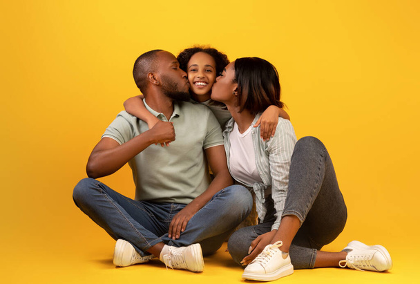 Conceito de valores familiares. Jovens pais africanos americanos beijando bochechas de sua filha, sentados em fundo amarelo - Foto, Imagem