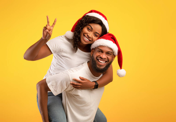 Emocionada pareja afroamericana en sombreros de Santa Divirtiéndose, chico dando a su novia paseo a cuestas sobre fondo amarillo - Foto, Imagen