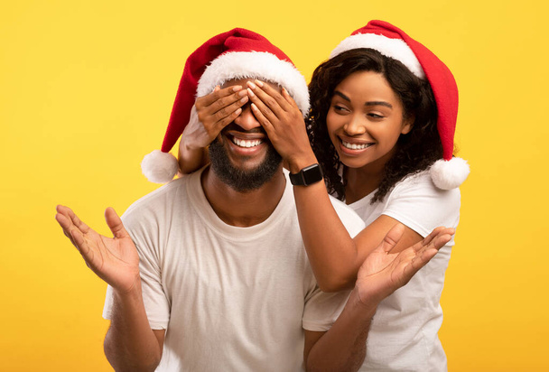 Beste cadeau voor Kerstmis. gelukkig afrikaanse amerikaanse dame staan achter de man en sluiten zijn ogen, geel studio achtergrond - Foto, afbeelding