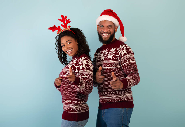 Hé, jij daar. Portret van gelukkig zwart paar in kerst truien wijzen naar camera over blauwe achtergrond - Foto, afbeelding