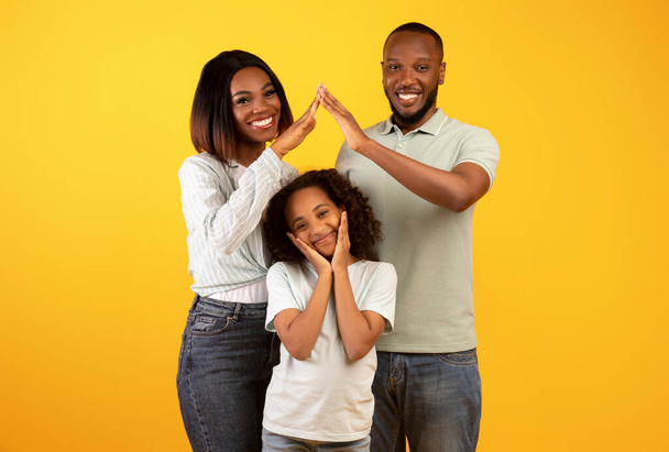Family care, protection and insurance concept. Young black parents making symbolic roof of hands above little daughter - Photo, Image