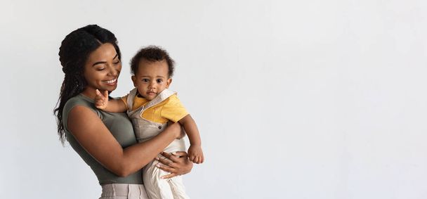 Moederschap concept. Glimlachende jonge zwarte moeder houden aanbiddelijke zuigeling zoon op armen - Foto, afbeelding