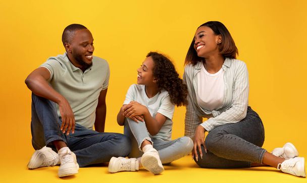 Concepto de comunicación. Felices padres negros y su hija hablando, sentados en el suelo de fondo amarillo - Foto, imagen