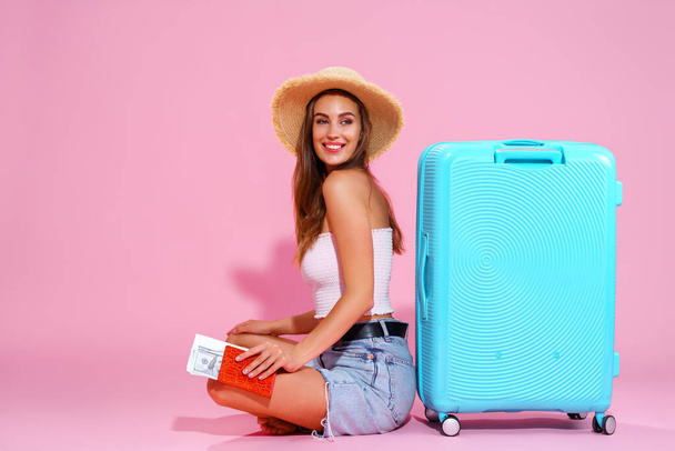 Smiling girl with a tickets are going to travel. Sitting near suitcase in shorts top and straw hat. Pink background  - Zdjęcie, obraz