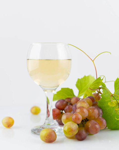White wine in a transparent glass on a white mirrored table. A bunch of grapes and a green vine in the background - 写真・画像