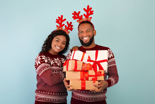 Yünlü kazak ve geyik boynuzu giyen mutlu siyah çift, mavi arka planda bir yığın Noel hediyesi tutan adam. - Fotoğraf, Görsel