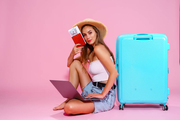  Smiling girl with laptop tickets money and passport are going to travel. Sitting near suitcase in shorts white top and straw hat. Pink background. looks at the camera - Фото, изображение