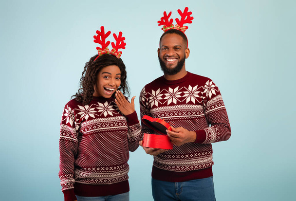 Excited black man giving wife Christmas gift, happy woman and her husband wearing deer horns, blue background - Photo, Image