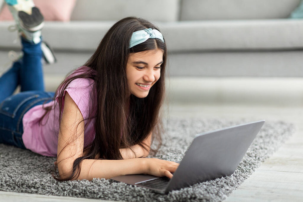 Concetto di apprendimento a distanza. Gioiosa ragazza adolescente che studia online, sdraiata su un tappeto morbido con laptop al chiuso - Foto, immagini