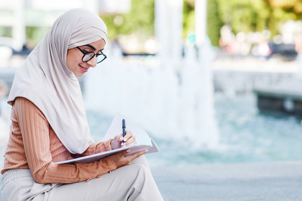 Cheerful cute young middle eastern female in hijab writing in notebook outdoor - Photo, Image
