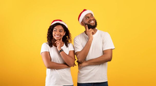 Мечтательные африканские супруги в шляпах Санта-Клауса держат пальцы на подбородке и думают о жёлтом фоне студии - Фото, изображение