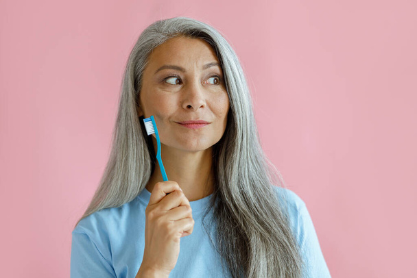 Positive thoughtful middle aged Asian woman holds toothbrush in studio - Foto, immagini