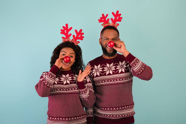 Weihnachtsspaß. Verspieltes schwarzes Paar mit Christbaumkugeln vor der Nase und Hirschhörnern vor blauem Hintergrund - Foto, Bild