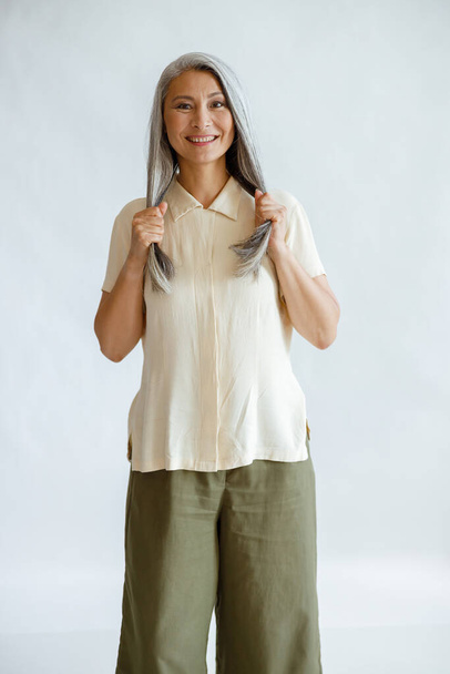 Улыбающаяся азиатка средних лет держит здоровые седые волосы на светлом фоне - Фото, изображение