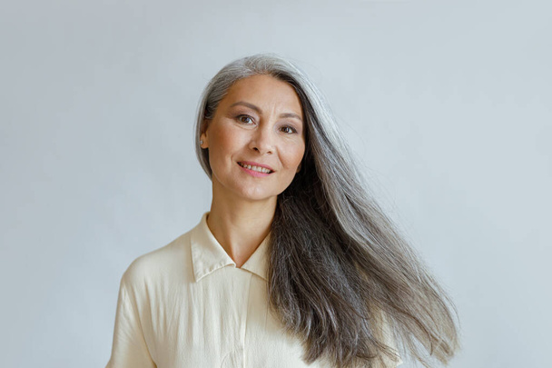 Sorrindo mulher asiática de meia idade com cabelos grisalhos fluindo fica no fundo claro - Foto, Imagem