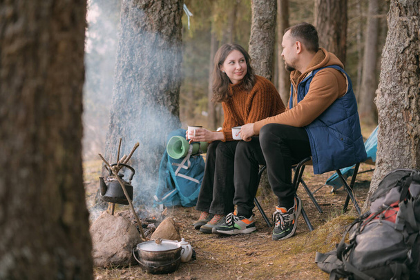 Family couple is resting on the shore of the lake with a tent. Fashing camp with a fire in the forest. A man and woman are shaking silence by fire with hot drink circles - Photo, Image